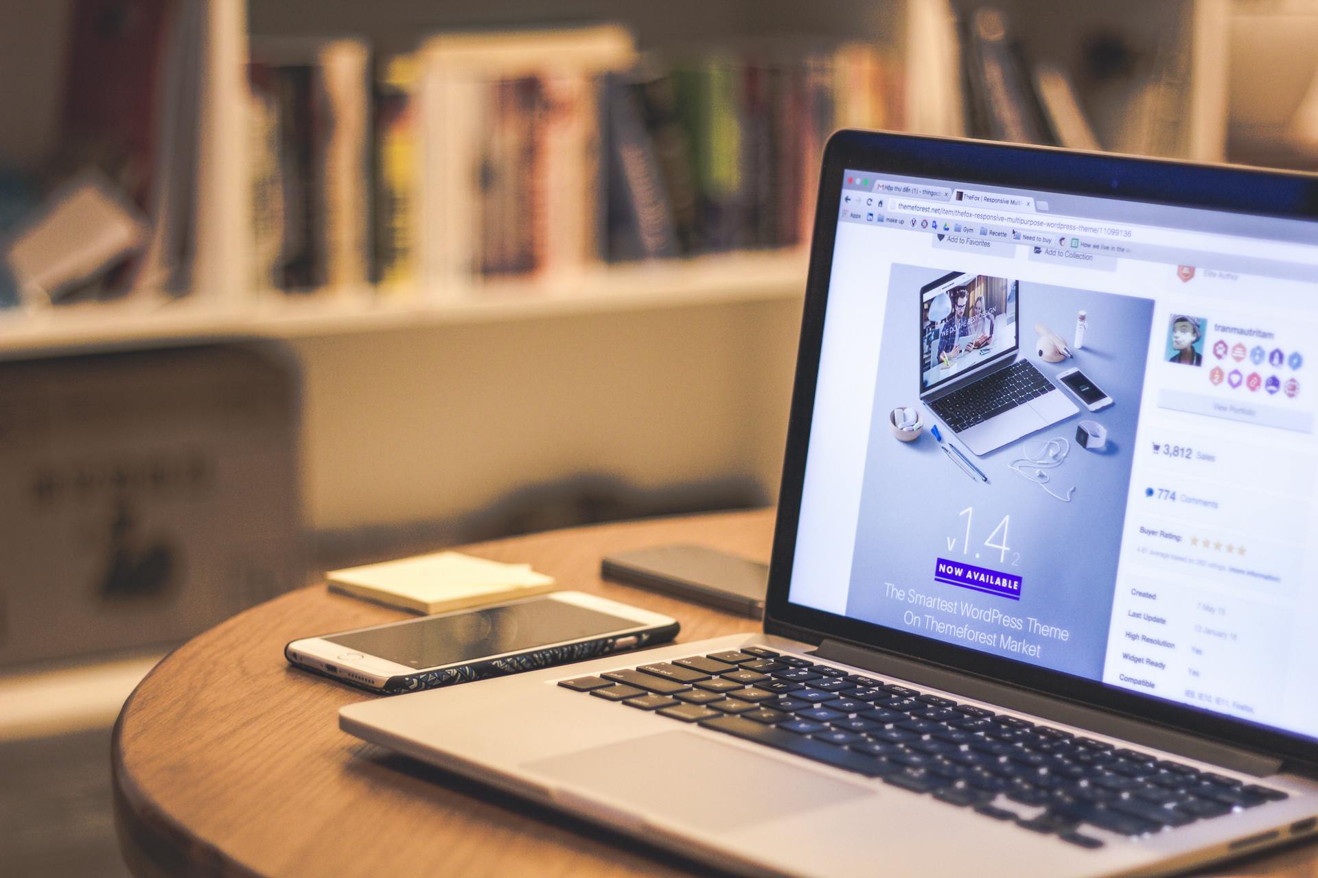 Laptop showing a website on a table with phones and a bookshelf behind