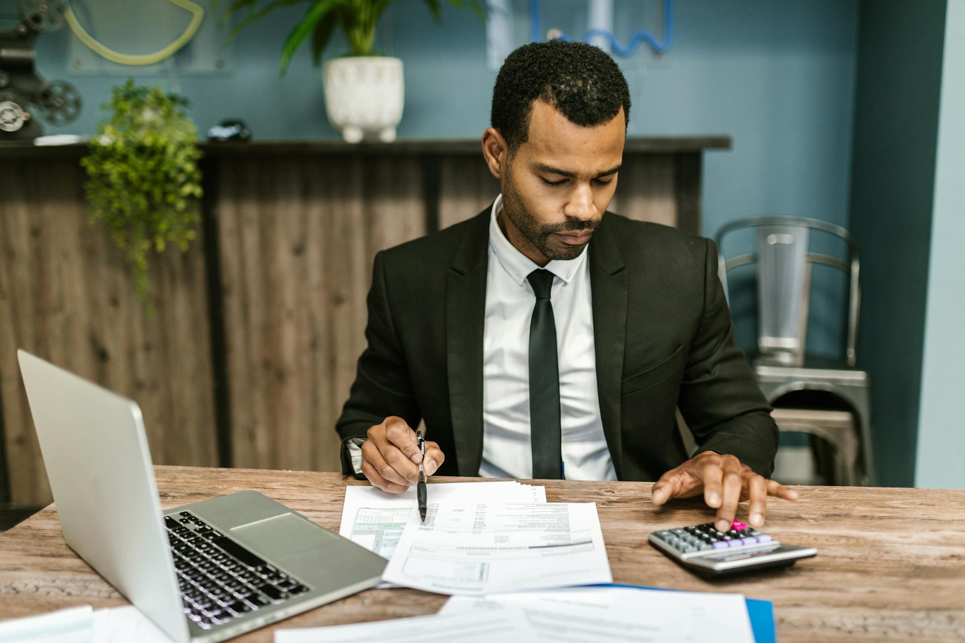 Man in a suit working through paperwork with a calculator and laptop